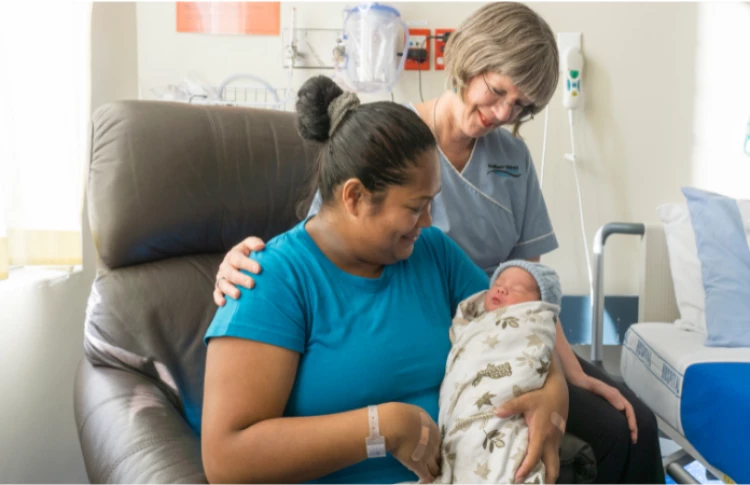 A hospital midwife sits supportively next to a mum carrying her newborn baby.